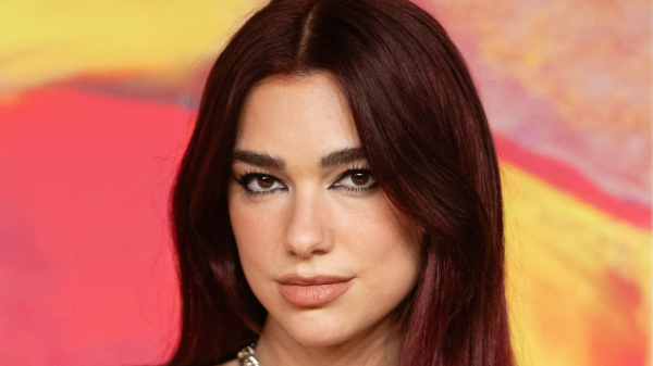 Dua Lipa poses at an event in a black leather dress. Her hair is dark red. She wears a silver necklace.