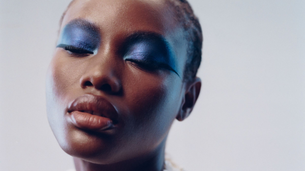 model wearing blue eyeshadow posing with her eyes closed wearing a white top with flower appliqus in front of a blue...